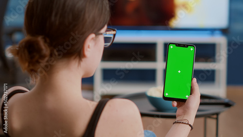 Closeup of young woman holding vertical smartphone with green screen watching social media content in home living room. Girl using touchscreen mobile phone with chroma key looking at screen.