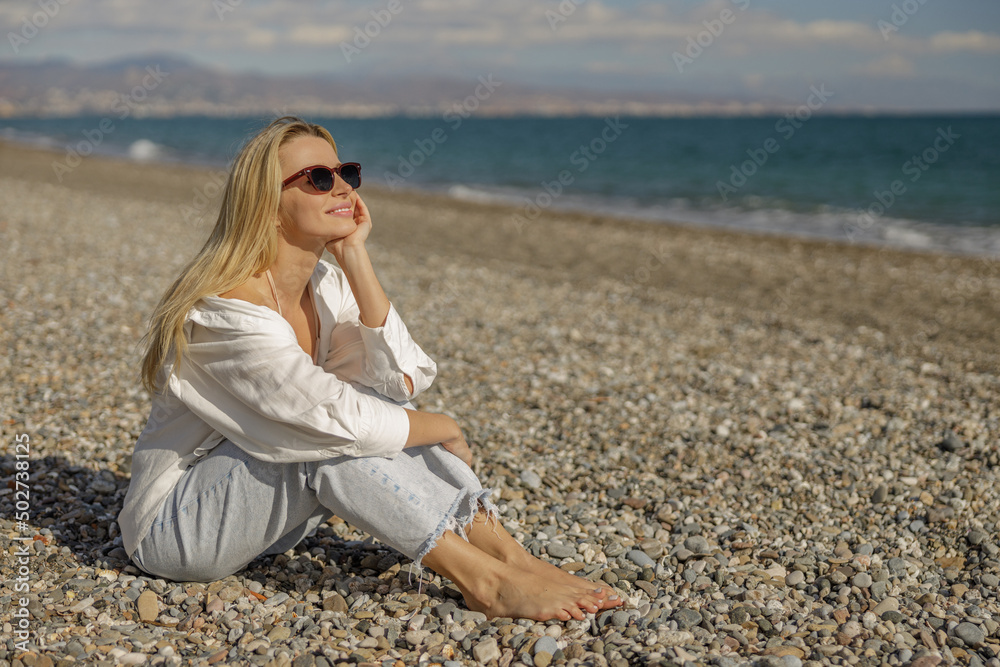 Dreamy woman in casual wear and sunglasses enjoying summertime, sitting barefoot on sea coast