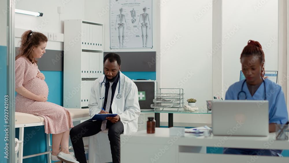 African american doctor writing checkup report on files to help patient with pregnancy belly. Male obstetrician giving maternity advice to expectant woman at consultation appointment.