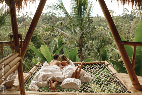 Happy travel couple on hammock balcony of bamboo tree house with jungle nature view. Vacation in beaitiful hidden place, honeymoon on Bali island photo