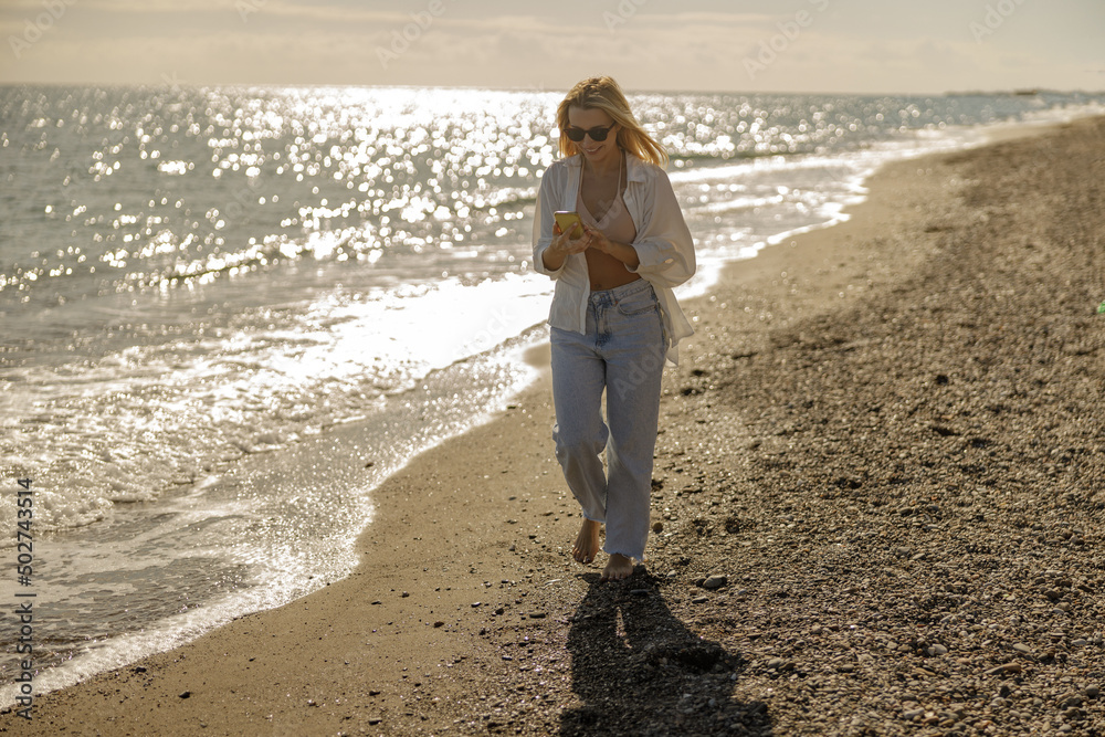 Full length of smiling woman chatting using smartphone, walking along the sea coast at sunset