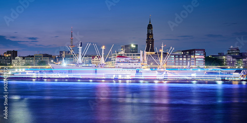 Fotografia Historic Cargo Vessel Cap San Diego in City of Hamburg during night time with blue colored light stripes from a tourism boat on the river Elbe