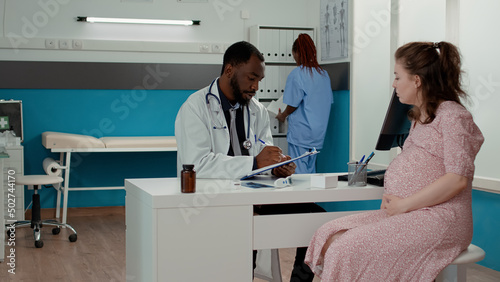 Physician consulting pregnant person and taking notes on checkup documents to give medical advice in cabinet. Male doctor having pregnancy discussion with patient expecting child.