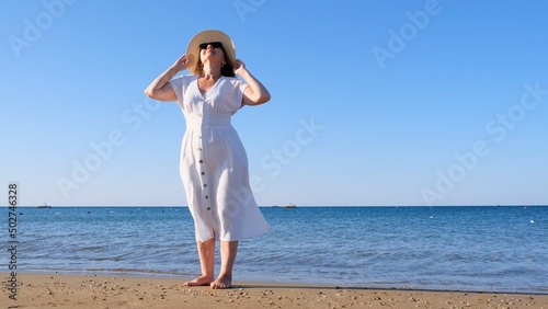 mature woman in sunglasses in a straw hat and white dress walks along the blue sea coast on a sunny summer day, enjoying freedom and relaxation. The concept of a senior citizen everyday life