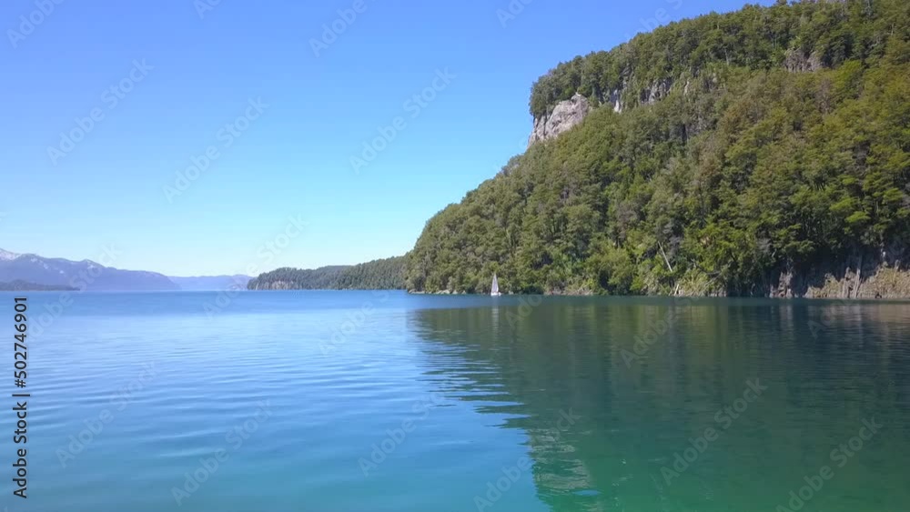 image that travels very close to the water until you get to see a sailboat up close sailing a lake with a mountain in the background. image that travels very close to the water until you get to see a sailboat up close sailing a lake with a mountain in the background.
