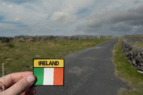 Tourist holding badge with Irish flag and sign Ireland in focus. Small country road and blue cloudy sky out of focus in the background. Travel and tourism concept. Irish landscape. photo