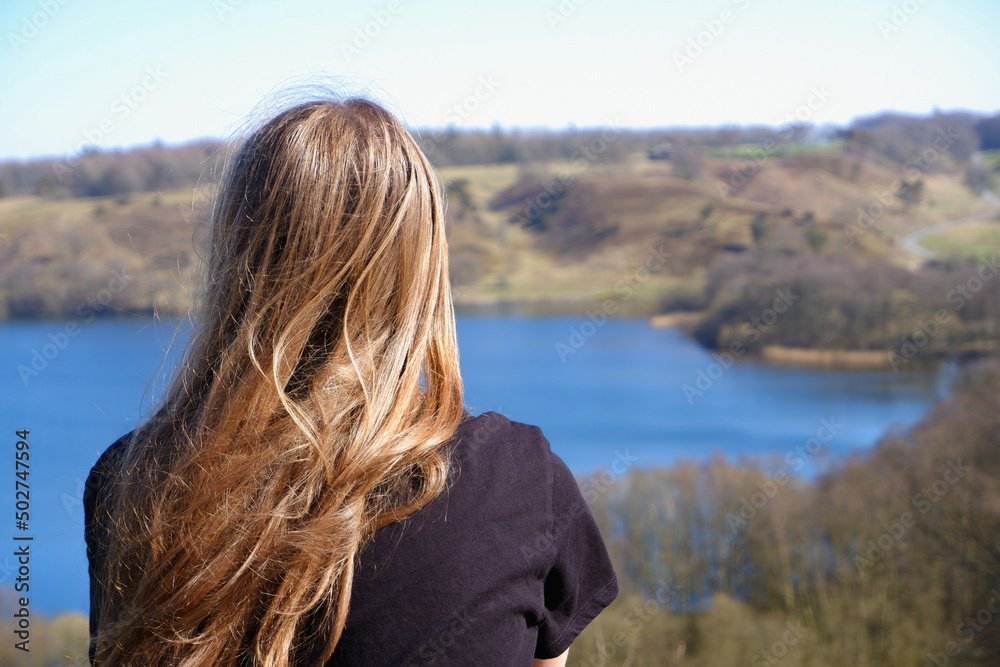 A girl is resting on a clear day on the shore of a blue lake with wooded hilly shores in early spring. View from the back. A girl is resting on a clear day on the shore of a blue lake with wooded hilly shores in early spring. View from the back.