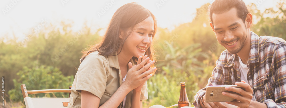 Fototapeta premium Happy, Cheerful asian young woman, girl and man holding, using smartphone sitting on chair outside tent. Adventure couple love camping in forest. Eco activity, lifestyle nature on holiday concept.