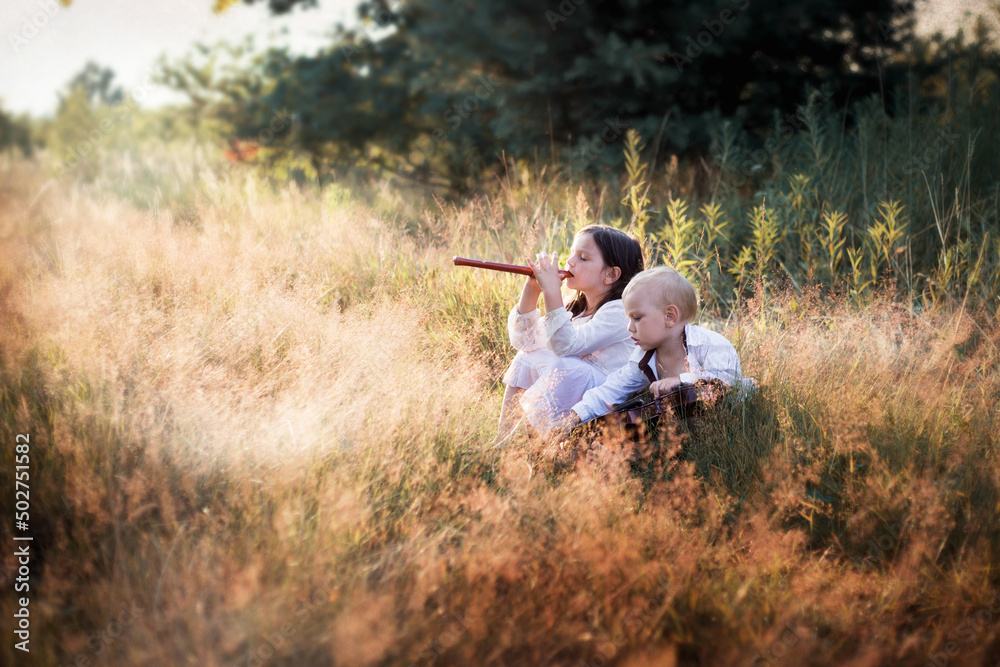 Dzieci grają na instrumentach siedząc na łące w wysokiej trawie, dziewczynka gra na flecie - obrazy, fototapety, plakaty 