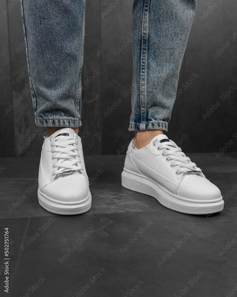 Close-up photo of men's white sneakers with jeans. White sneakers on his feet Close-up photo of men's white sneakers with jeans. White sneakers on his feet