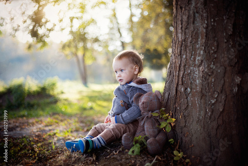 Malutki chłopiec siedzi pod drzewem w parku razem ze swoim misiem i patrzy na wodę