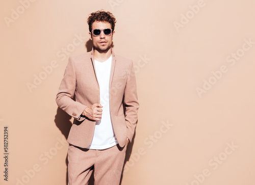 Portrait of handsome confident stylish hipster lambersexual model. Sexy modern man dressed in elegant beige suit. Fashion male with curly hairstyle posing in studio. Isolated. In sunglasses © halayalex