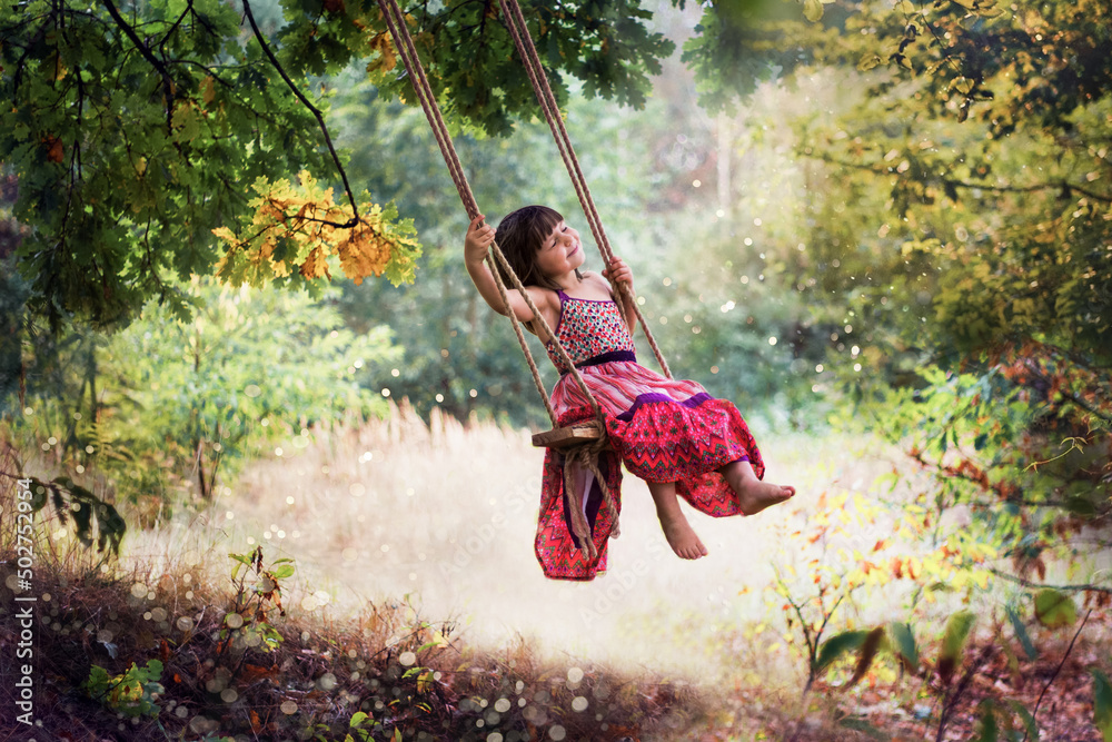 Dziewczynka w letniej sukience huśta się na huśtawce powieszonej na drzewie, śmieje się i śpiewa piosenki wymachując nogami - obrazy, fototapety, plakaty 