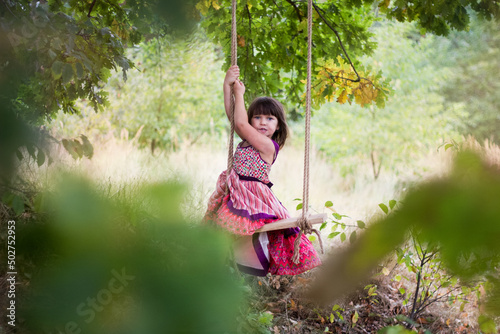 Dziewczynka w letniej sukience huśta się na huśtawce powieszonej na drzewie, śmieje się i śpiewa piosenki wymachując nogami