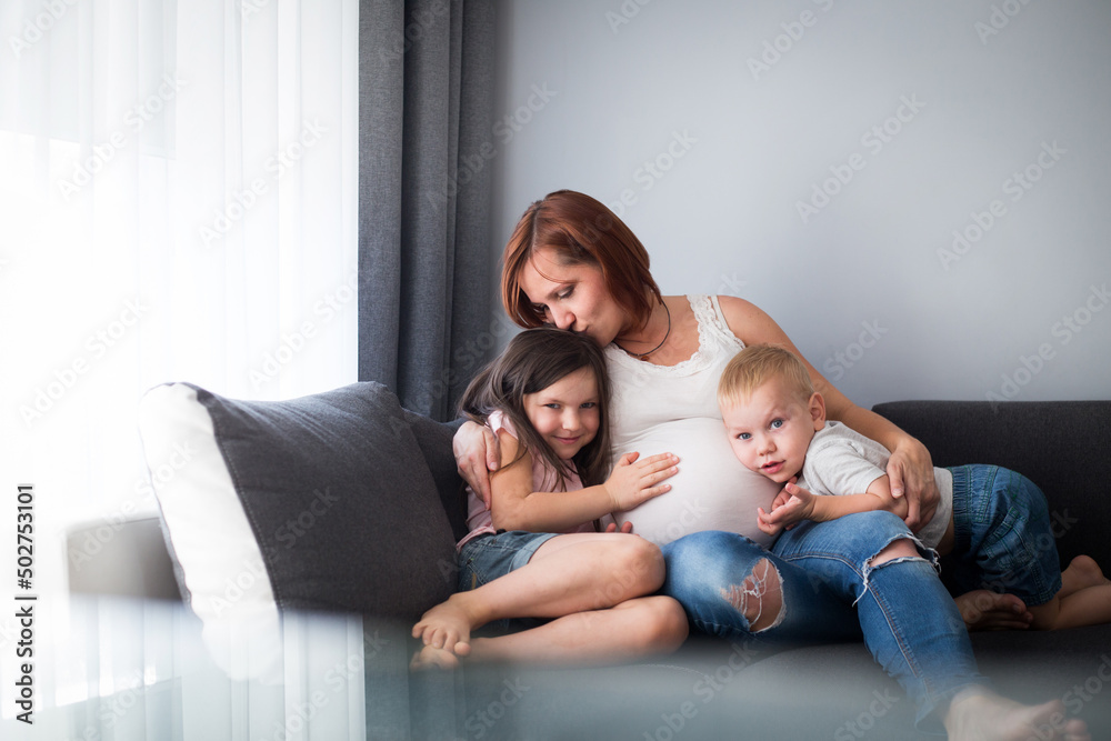 Kobieta w ciąży ze swoimi dziećmi siedzi w domu na kanapie, mama przytula w objęciach swoje dzieci - obrazy, fototapety, plakaty 