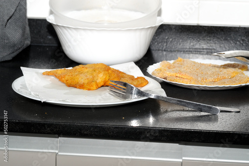 Close-up of breaded chicken breast meat, fried and crispy, on a white plate with forks and next to unfried breaded chicken fillets. Placed on the black kitchen countertop. Photo with space to copy photo