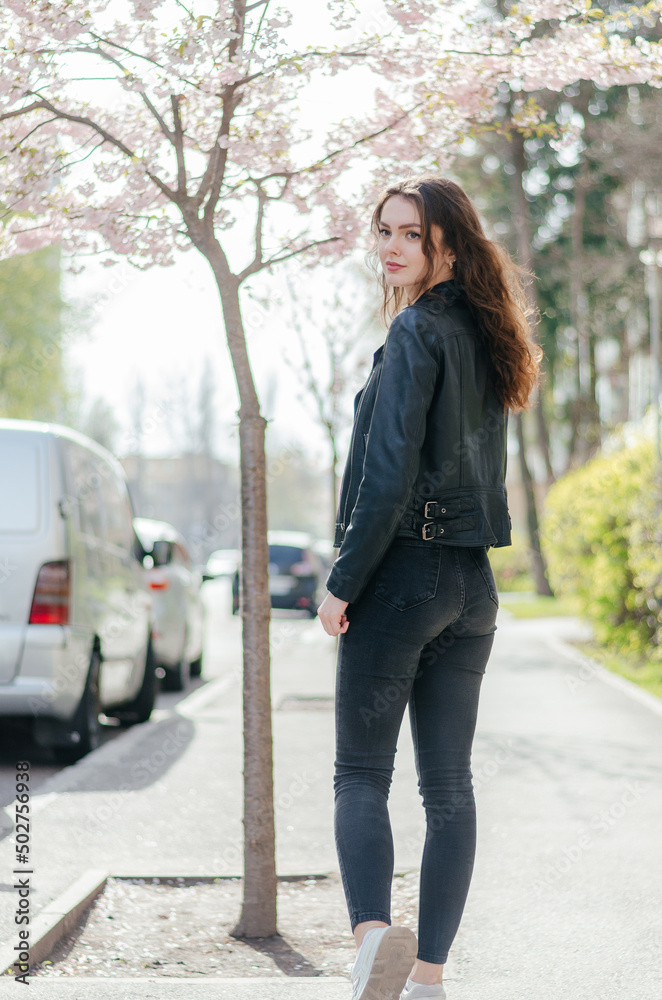 girl in black jeans and a leather jacket walks on the sidewalk near the sakura tree
