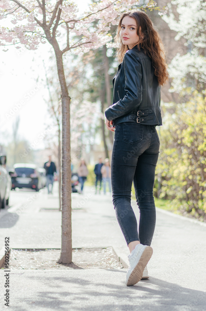 girl in black jeans and a leather jacket walks on the sidewalk near the sakura tree girl in black jeans and a leather jacket walks on the sidewalk near the sakura tree