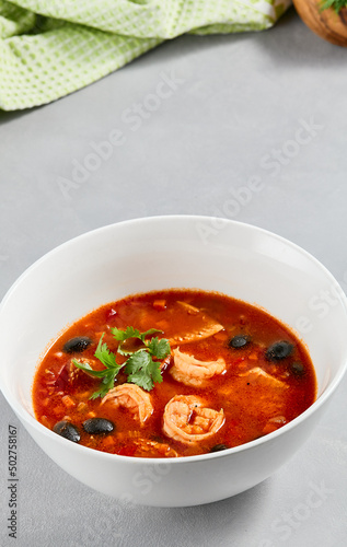 Fish tomato soup with shrimps and black olives. Seafood soup with prawns in white ceramic bowl on concrete background. Fish dish with seafood. Tomato soup in minimal composition.