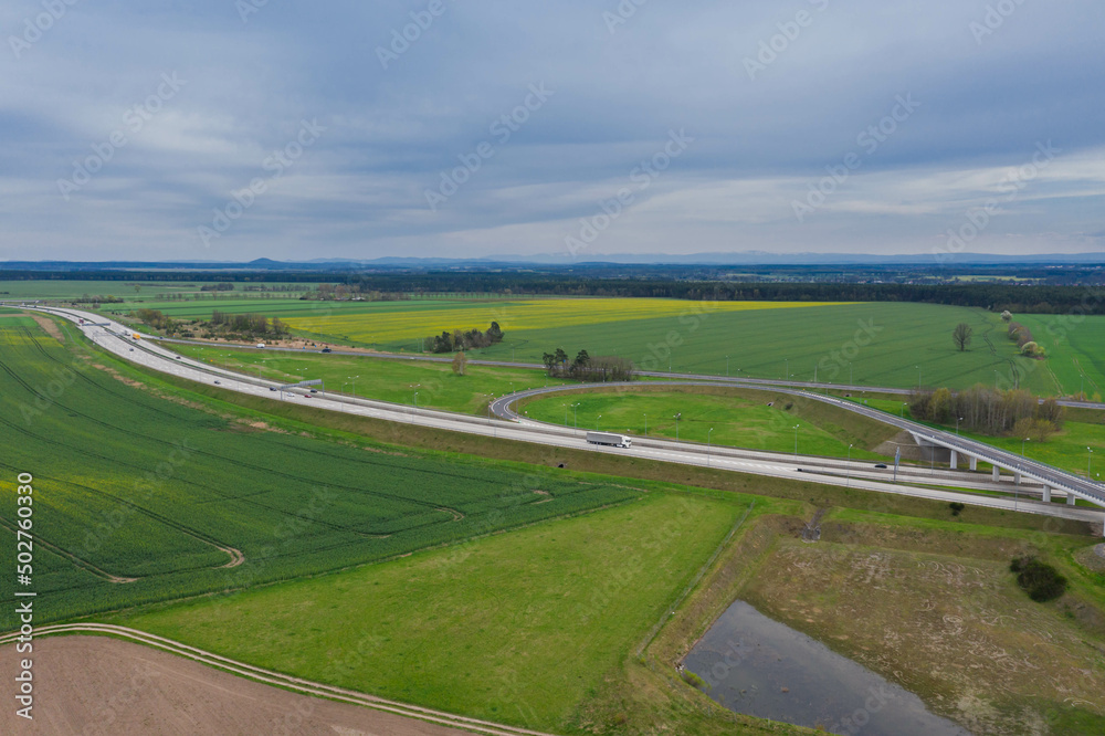 Rozległa równina, zielone pola uprawne i łąki. W centrum kadru widoczny jest duży węzeł komunikacyjny, rozwidlenie dróg szybkiego ruchu. Widok z dużej wysokości. Zdjęcie z drona.