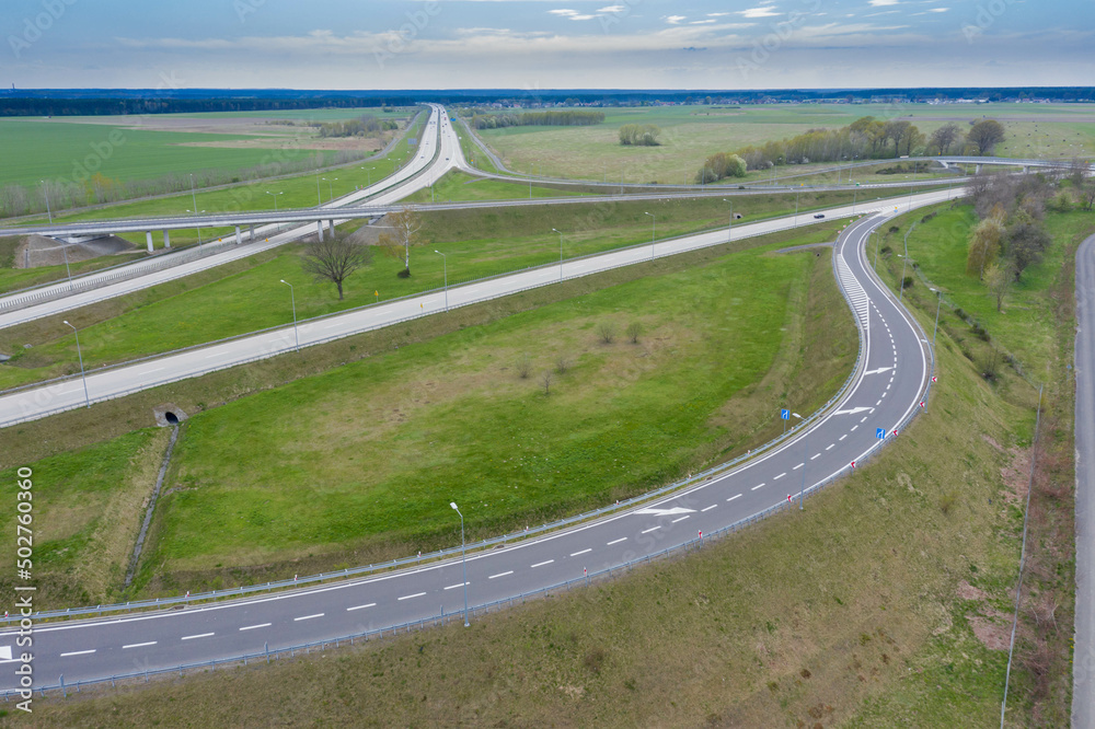 Rozległa równina, zielone pola uprawne i łąki. W centrum kadru widoczny jest duży węzeł komunikacyjny, rozwidlenie dróg szybkiego ruchu. Widok z dużej wysokości. Zdjęcie z drona.