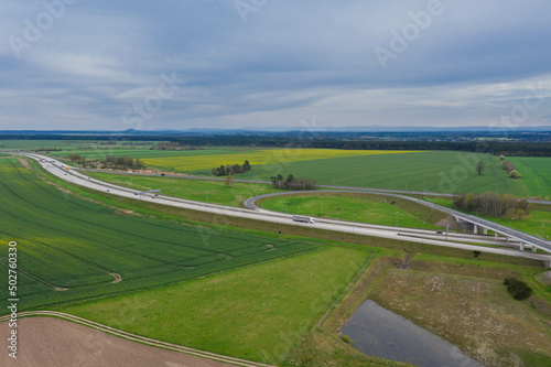 Rozległa równina, zielone pola uprawne i łąki. W centrum kadru widoczny jest duży węzeł komunikacyjny, rozwidlenie dróg szybkiego ruchu. Widok z dużej wysokości. Zdjęcie z drona.