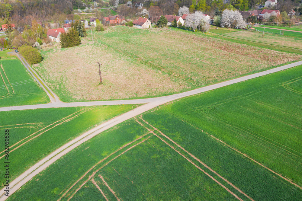 Droga polna przebiegająca przez pola i łąki widziana z wysokości. Zdjęcie z drona. W oddali widać zabudowania pobliskiej wioski. - obrazy, fototapety, plakaty 