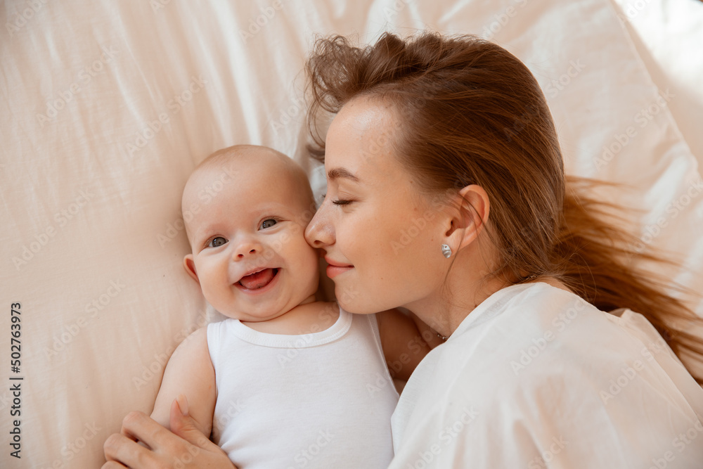 Happy young mother with baby at home in white room, mom and baby are lying, kissing the baby at home on the bed in the bedroom Happy young mother with baby at home in white room, mom and baby are lying, kissing the baby at home on the bed in the bedroom