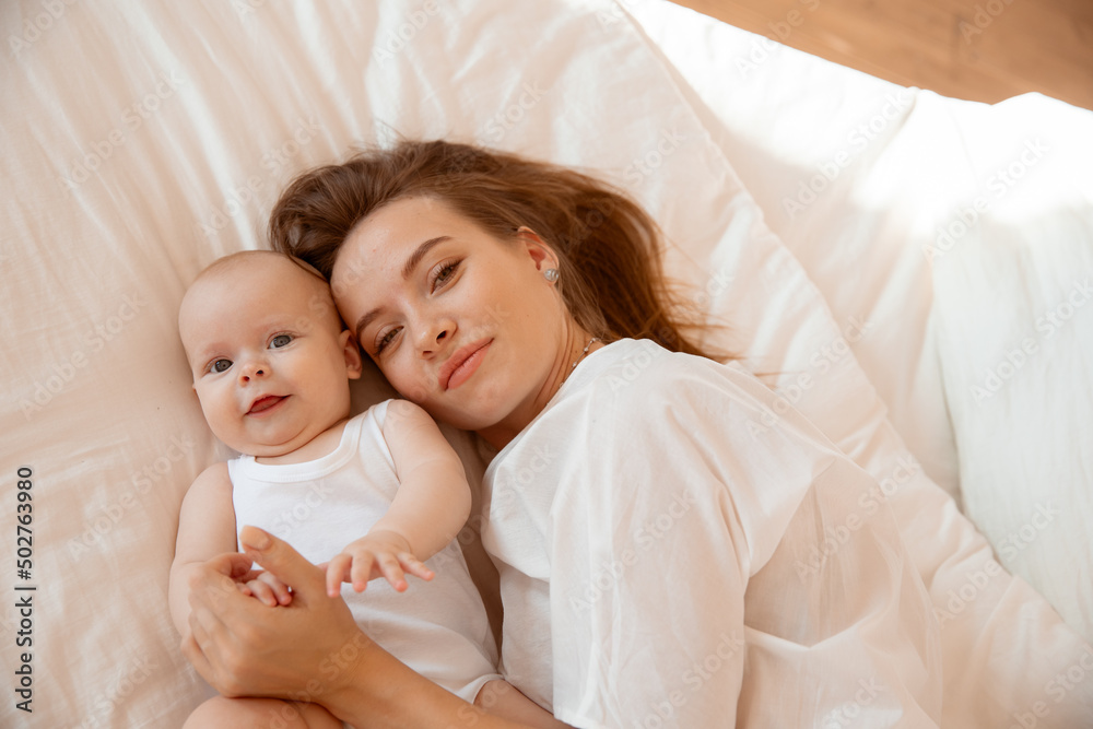 Happy young mother with baby at home in white room, mom and baby are lying, kissing the baby at home on the bed in the bedroom Happy young mother with baby at home in white room, mom and baby are lying, kissing the baby at home on the bed in the bedroom