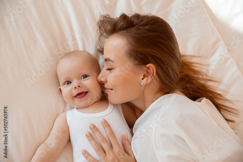 Happy young mother with baby at home in white room, mom and baby are lying, kissing the baby at home on the bed in the bedroom