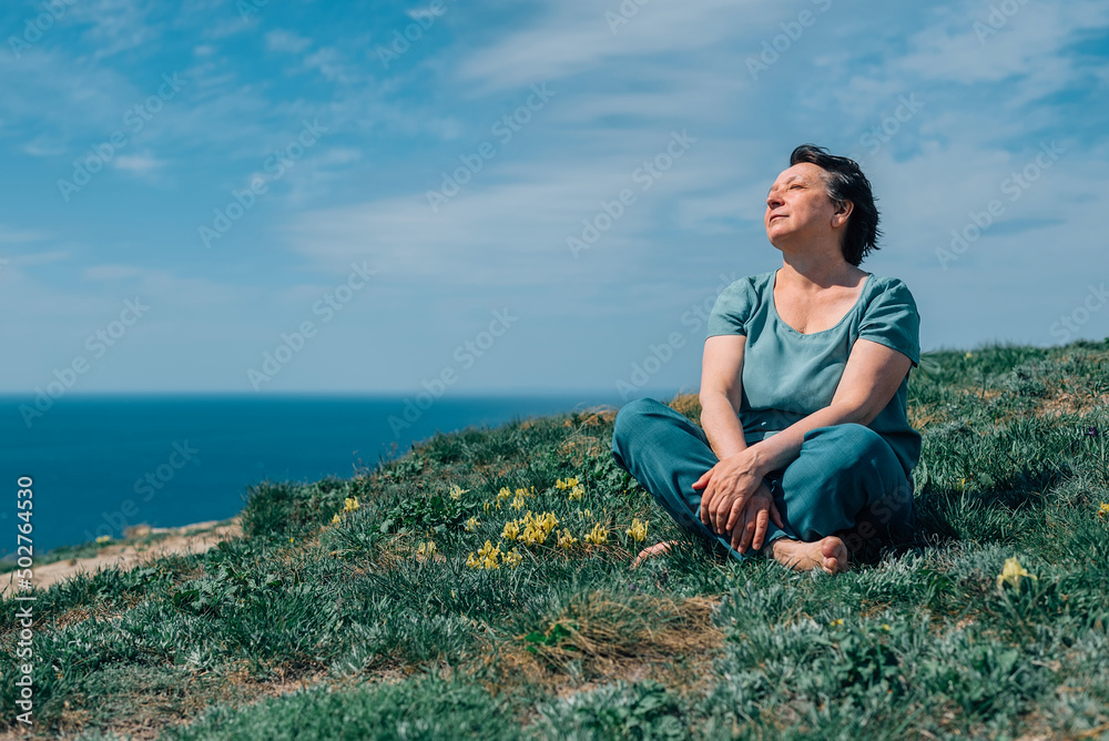 an adult woman, pensioner sits on the grass, legs crossed in a lotus position barefoot with a smile on her face on a sunny day on a mountain against the backdrop of the sea in summer, spring
