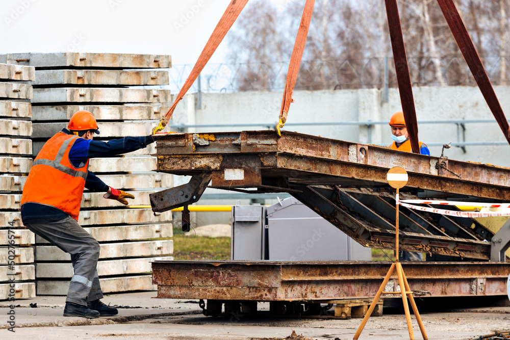 Naklejka premium Slinger in helmet and vest controls unloading of metal structures on construction site. White handyman unloads load.