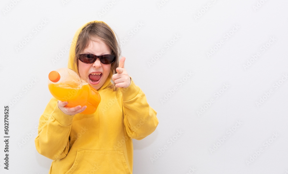 Caucasian girl 6 years old, in a yellow sweatshirt with a hood on a gray background. The child smiles and poses for the camera, holding a bottle of sweet soda in his hands. Caucasian girl 6 years old, in a yellow sweatshirt with a hood on a gray background. The child smiles and poses for the camera, holding a bottle of sweet soda in his hands.