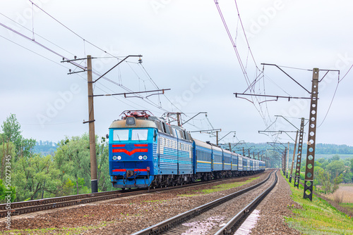 A powerful dual-system electric locomotive pulls a long intercity passenger train to the railway station. Spring cloudy weather.
