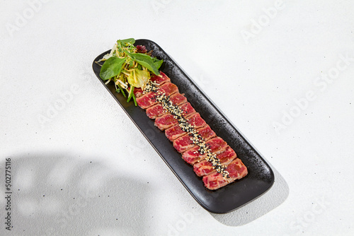 Beef tataki with green salad in black plate on white table with hard shadow. Meat carpaccio in asian style. Traditional asian food from sliced meat - tataki in minimal design with sun light shadow. photo