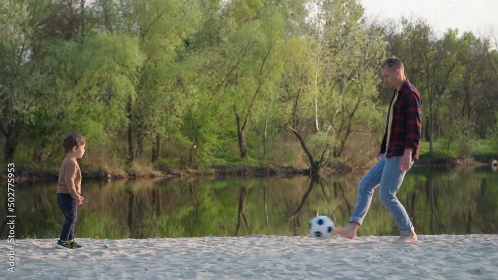 Father Plays Soccer with Son on Sand. Slow Motion. Young Man And Little Boy Playing With Black and White Football Ball Outdoors Near River Father Plays Soccer with Son on Sand. Slow Motion. Young Man And Little Boy Playing With Black and White Football Ball Outdoors Near River