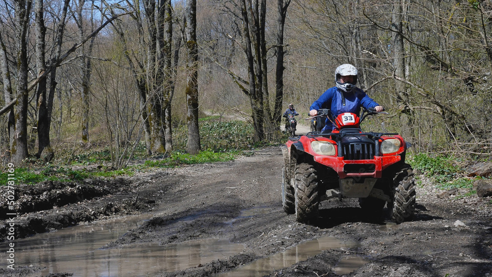 Fototapeta premium A red quad bike with a girl behind the wheel is about to drive through a puddle in the spring woods. Front view. Extreme type of outdoor activities. ATV riding.