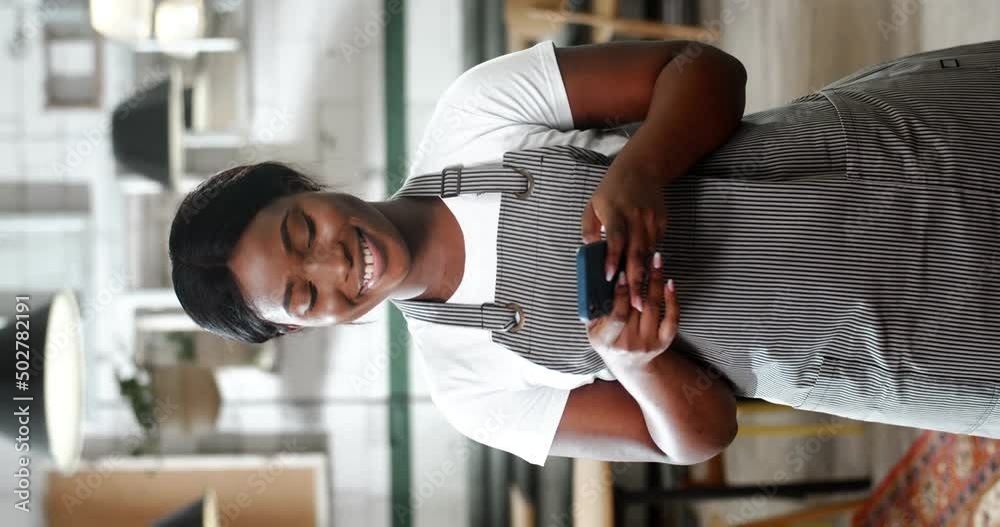 Vertical portrait of beautiful African American young woman waiter in apron standing cafe kitchen, using smartphone and looking at camera with smile. Female chef of restaurant texting on mobile phone. Vertical portrait of beautiful African American young woman waiter in apron standing cafe kitchen, using smartphone and looking at camera with smile. Female chef of restaurant texting on mobile phone.