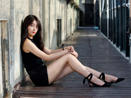 Graceful young woman with long black hair sitting on ground and leaning on the wall looking at camera in sunny day. Outdoor portrait of cool Chinese girl in sexy black sundress, summer concept.