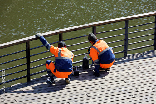 Photography Workers in uniform paints the metal fence on a lake embankment