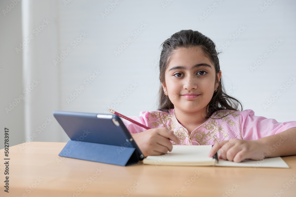 portrait cute Indian girl writing on notebook and online learning class from tablet, education concept portrait cute Indian girl writing on notebook and online learning class from tablet, education concept