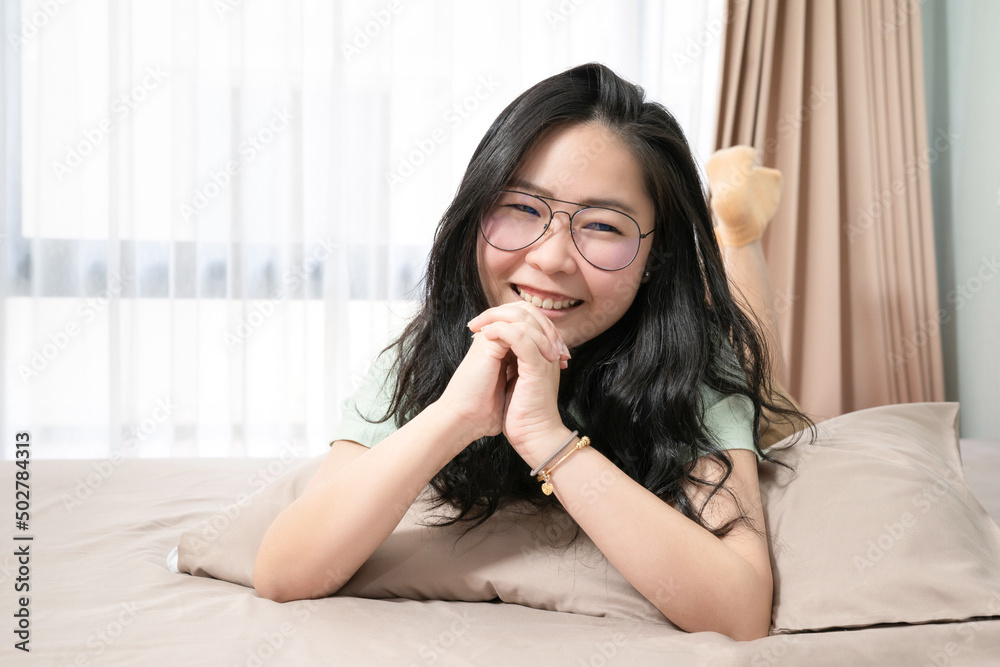 Beautiful Asian woman hold her hands together while lie on her stomach on bed in bedroom with a pastel green - brown color theme. Beautiful Asian woman hold her hands together while lie on her stomach on bed in bedroom with a pastel green - brown color theme.