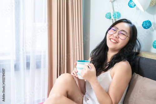 Asian Glasses Woman holds a cup of coffee and relax on the bed in the morning at her bedroom. photo