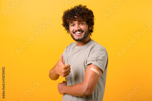 Smiling young curly Indian guy showing arm with band-aid after vaccine injection Fototapete
