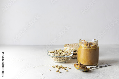 A transparent jar of sunflower seed oil on a light table. Seeds in a glass vase and bread in the background. Spoon with butter on the table. Light background, space for text