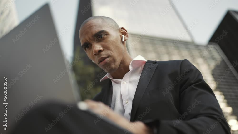 Displeased African businessman working on laptop and talking in headphones outdoors Displeased African businessman working on laptop and talking in headphones outdoors