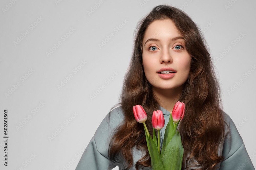 Young woman with tulips on a gray background. Blue eyes, long hair and surprise on her face. Young woman with tulips on a gray background. Blue eyes, long hair and surprise on her face.