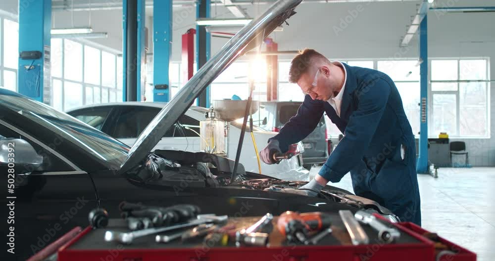 Caucasian male mechanic in uniform and goggles repairing car motor in auto service. Young man engineer fixing engine of vehicle in big garage. Maintaining concept. Repairment work.
