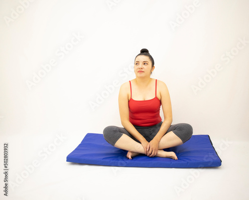 Beautiful white woman in red and gray sportswear, sitting and resting on a blue mat after exercising, on white background.