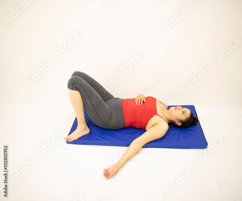 beautiful athletic white oriental woman resting in gray sportswear and red blouse with her legs retracted, on blue mat, on white background